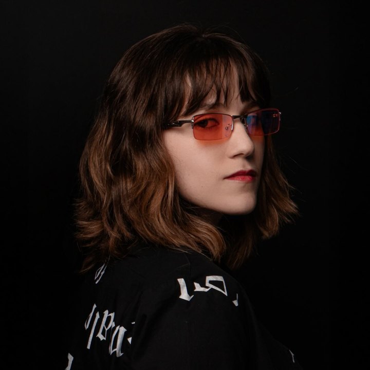 Isabela Angelo, de óculos com lentes coloridas, sobre fundo escuro